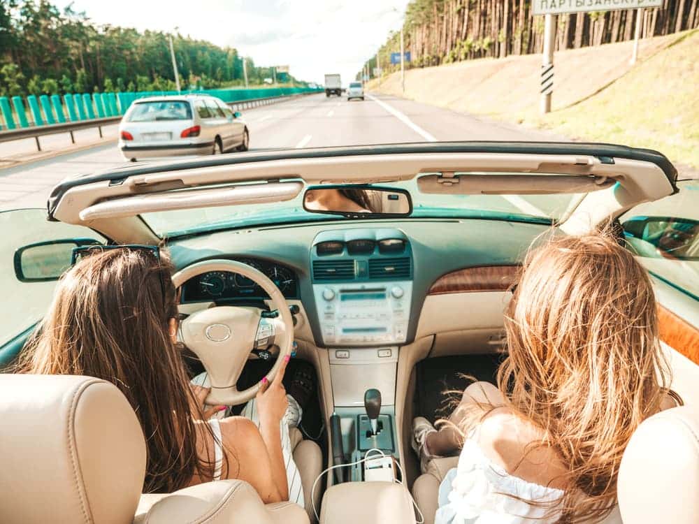 A depiction of two youthful and cheerful hipster women in a convertible car. These optimistic models are enjoying themselves, clad in sunglasses, as they ride along.