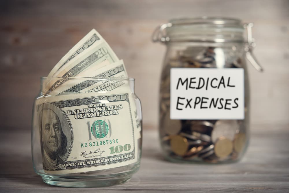 In a vintage-toned wooden background with dramatic lighting, there's a glass jar filled with dollars and coins labeled "Medical Expenses," illustrating a financial concept.