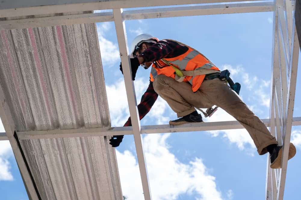 A roofer wearing protective workwear and gloves, along with a construction worker in a safety harness, working at a high level on a construction site installing something.