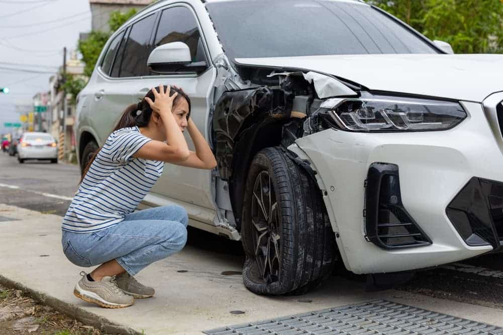 A woman squatting next to a damaged white car, holding her head in distress.