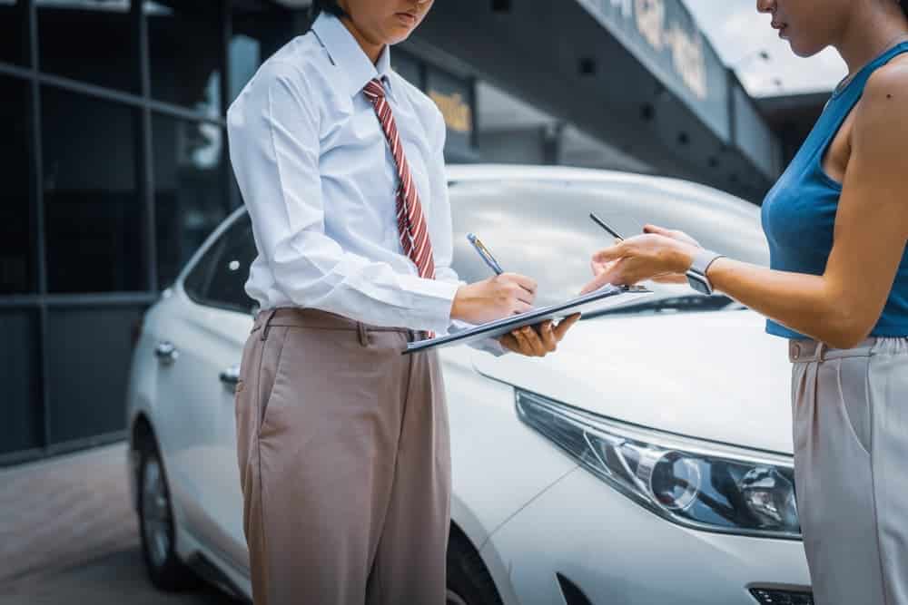 Two people filling out paperwork in front of a white car, focusing on documents.