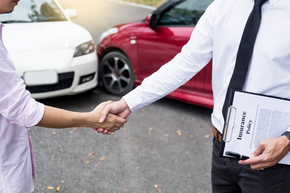 Two men shake hands in front of a white and a red car after an accident, with one holding an insurance policy clipboard.