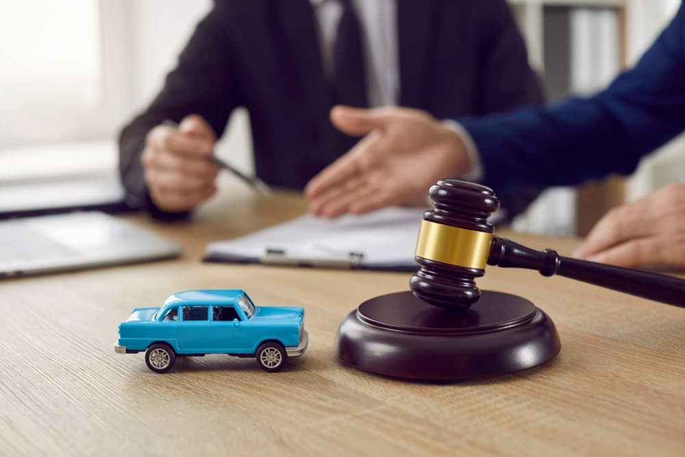 A gavel and toy car on a desk, with two people discussing paperwork in the background.