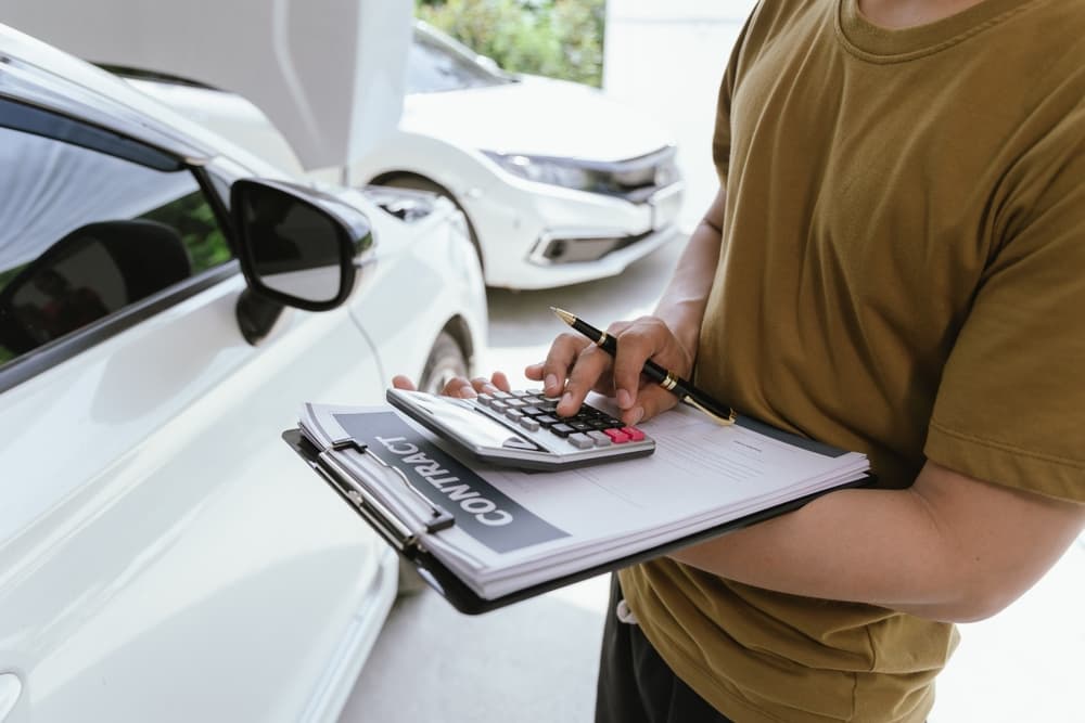 A young man holds a calculator and a file to assess insurance policy limits, standing next to a dented car after an accident.